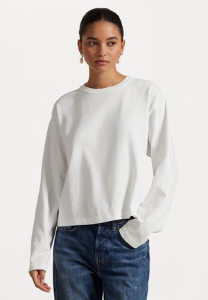 Hvit langermet cropped genser laget av myk bomull, med rund hals og avslappet passform. Kombinert med blå denimjeans.
