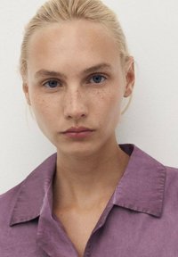 Portrait of a person with light blonde hair and blue eyes, wearing a lavender collared shirt with a textured fabric.