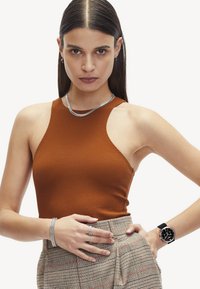 Brauner ärmelloser Top mit strukturiertem Finish, kombiniert mit einem karierten High-Waist-Rock. Die Accessoires umfassen eine silberne Kettenhalskette, ein Armband und eine Uhr.