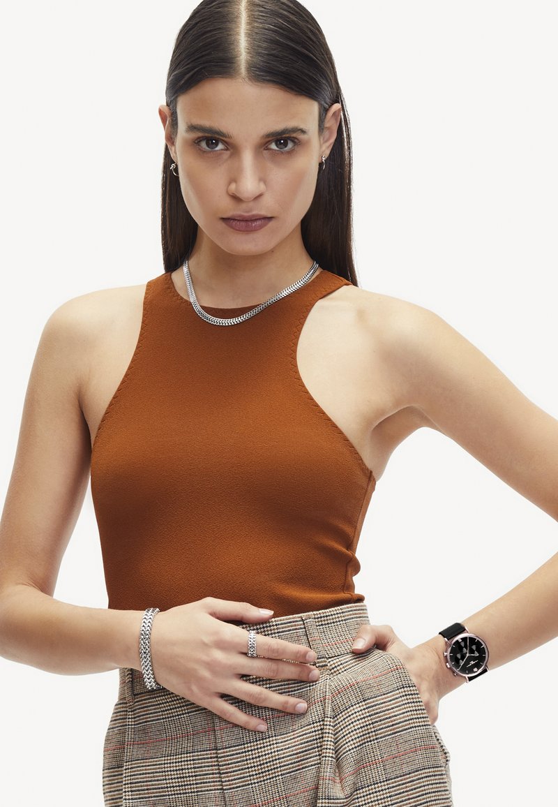 Brauner ärmelloser Top mit strukturiertem Finish, kombiniert mit einem karierten High-Waist-Rock. Die Accessoires umfassen eine silberne Kettenhalskette, ein Armband und eine Uhr.