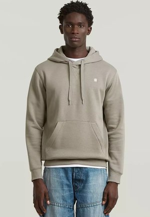 Jonge man draagt een lichtbeige hoodie met voorzak en blauwe spijkerbroek, staand tegen een effen lichtgrijze achtergrond.