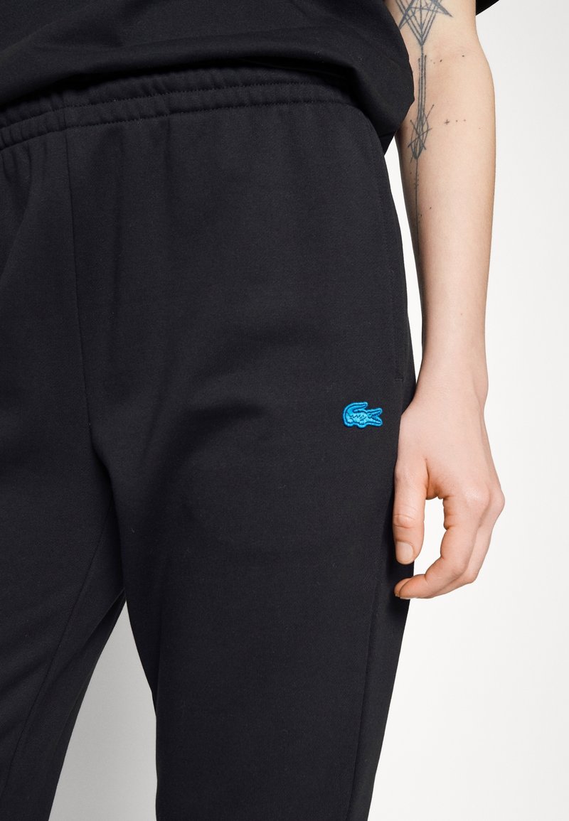 Czarne dresy wykonane z miękkiego materiału, z elastycznym paskiem w talii i bocznymi kieszeniami. Logo Lacoste jest wyszyte na niebiesko.