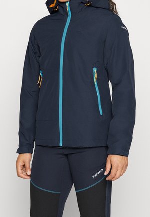 Osoba nosząca granatową kurtkę outdoor z turkusowymi zamkami i dopasowane granatowe spodnie sportowe z logo na udzie.