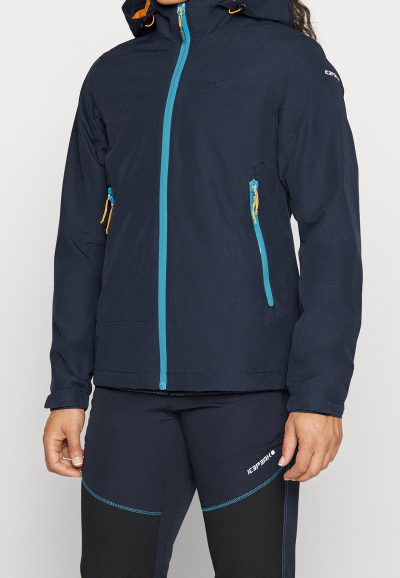 Personne portant une veste d’extérieur bleu marine avec des fermetures éclair turquoise et un pantalon de sport bleu marine assorti avec un logo sur la cuisse.