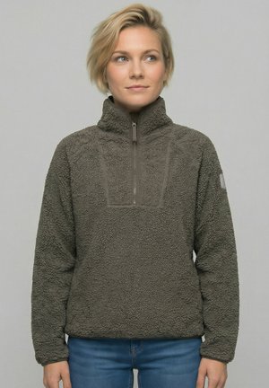 Ung person med kort blondt hår iført en grå fleece-pullover med lynlås foran, stående mod en ensfarvet baggrund.