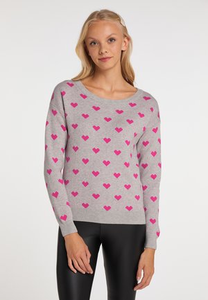 Junge Frau trägt einen grauen Pullover mit pinken Herzen und schwarze Hose, steht vor einem einfarbigen hellen Hintergrund.