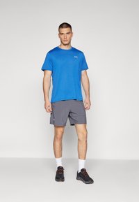 Conjunto deportivo para hombre que incluye una camiseta de manga corta azul y unos pantalones cortos grises, ambos con logotipos. Zapatillas de deporte negras con acentos naranjas y calcetines blancos.