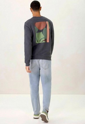 Grauer Sweatshirt mit einer großen abstrakten Grafik in Grün, Rot und Schwarz auf dem Rücken. Kombiniert mit hellblauen Jeans und grünen Sneakers.