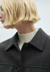 Grå mønstret uldfrakke med krave og knaplukning, båret over en cremefarvet ribbet turtleneck. Kort, blond hår synligt i kanten.