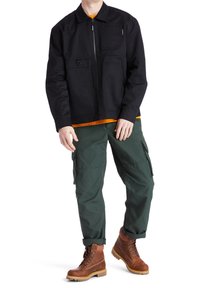 Černá zipová bunda s dvěma předními kapsami, nošena přes oranžové tričko. Zelené cargo kalhoty a hnědé šněrovací boty dokončují tento outfit.