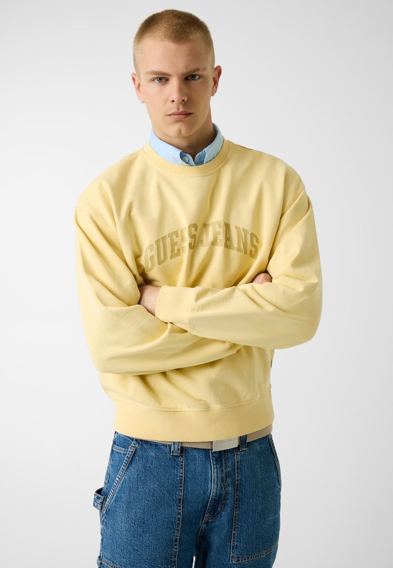 Jeune homme aux cheveux courts portant un sweat-shirt jaune GUESS JEANS par-dessus une chemise bleu clair et un jean en denim bleu, bras croisés.