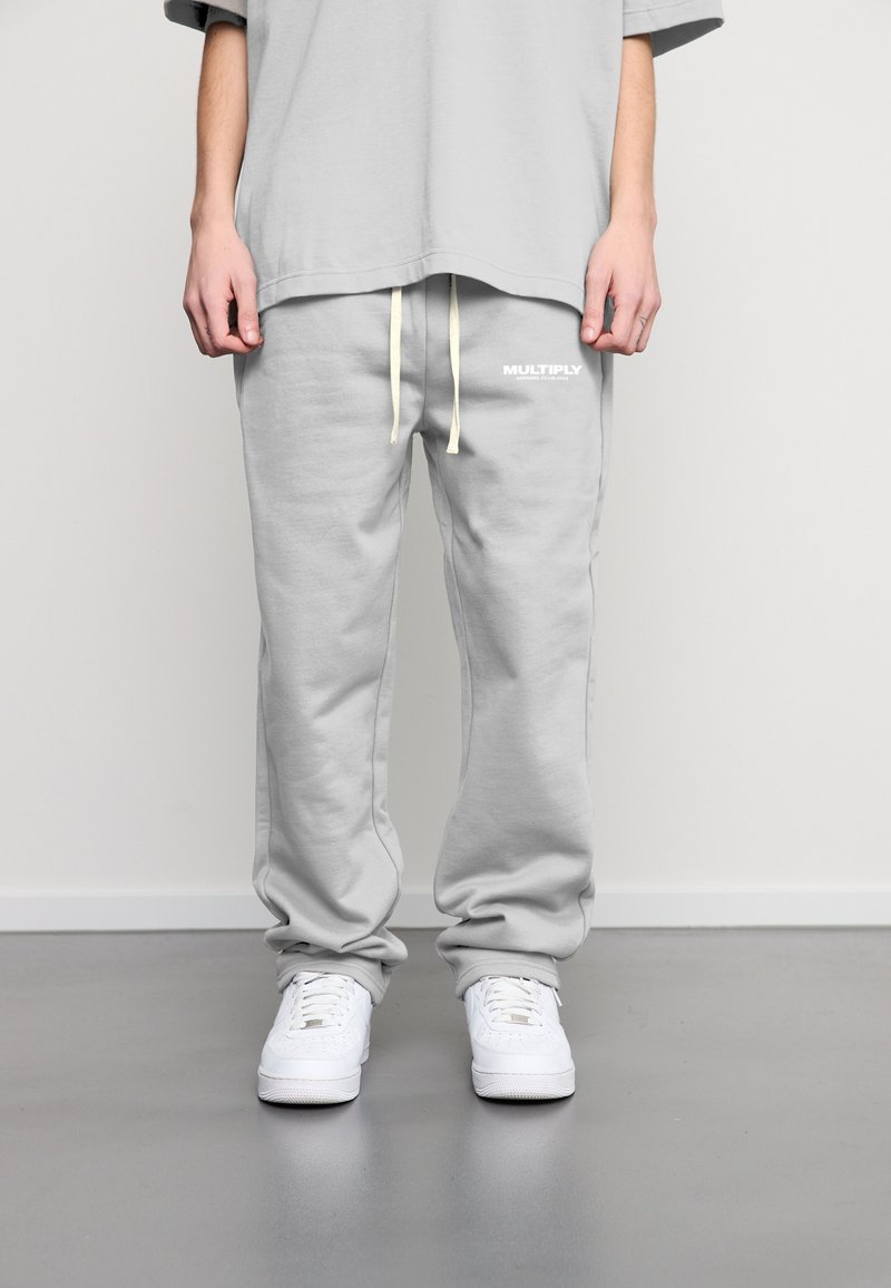 Grijze joggingbroek van zacht materiaal, met een losse fit, elastische tailleband met trekkoord en een wit "MULTIPLY" logo aan de linkerkant.