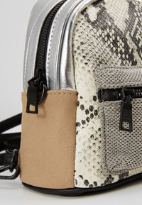 Gros plan sur un coin de sac à main texturé avec imprimé serpent, suède beige, garniture argentée, poche en tissu et tirettes de fermeture éclair noires.