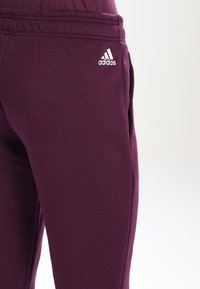 Lilla sweatpants lavet af blødt stof, med en tapered design, elastisk talje og hvid Adidas-logo på venstre side.