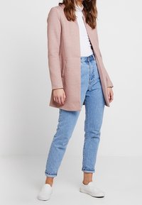 Cappotto lungo rosa chiaro, con apertura frontale e senza bottoni. Abbinato a jeans blu a vita alta e scarpe slip-on bianche. Texture liscia.