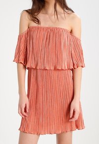 Robe plissée à épaules nues en corail, présentant un tissu texturé, une silhouette fluide et un ourlet évasé. Les manches courtes créent un effet superposé.