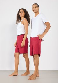 To modeller står side om side i uformell bekledning. Kvinnen har på seg en hvit tank topp og røde shorts, mens mannen har en hvit t-skjorte og røde shorts.