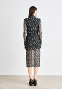 Black mesh dress with green eye pattern, long sleeves, and a mid-length cut. Features a fitted silhouette and layered design. Black high-heeled shoes.