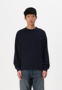 Μπλέ ναυτική μπλούζα μακρύ μανίκι φτιαγμένη από μαλακό ύφασμα, με τσέπη στο αριστερό μέρος του στήθους και άνετη εφαρμογή, σε συνδυασμό με φαρδιά τζιν παντελόνια.