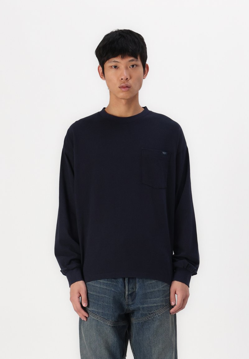 Μπλέ ναυτική μπλούζα μακρύ μανίκι φτιαγμένη από μαλακό ύφασμα, με τσέπη στο αριστερό μέρος του στήθους και άνετη εφαρμογή, σε συνδυασμό με φαρδιά τζιν παντελόνια.