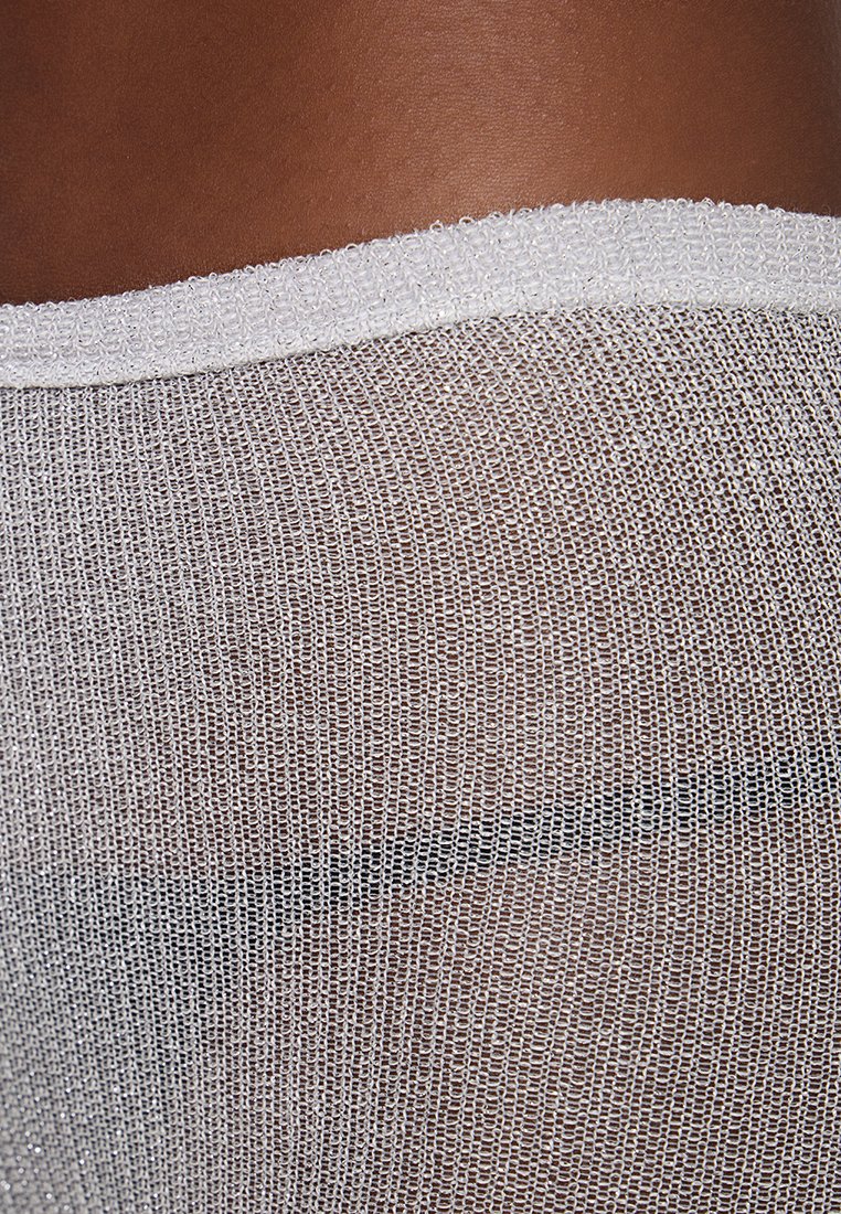 Przezroczysty, metaliczny, dziany materiał o teksturowanym wzorze w kolorze srebrnym. Krawędź zdobiona jest jednolitą białą lamówką. Tło w ciemnym odcieniu skóry.