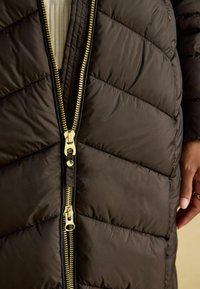 Hnedý nafukovací kabát s chevron vzorom, dvojitým zlatým zipsom a textúrovanou tkaninou. Exponované švy pridávajú hĺbku.