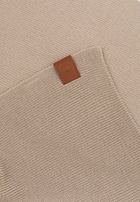 Beżowa dzianina z teksturowanym wzorem, z kwadratową brązową skórzaną metką z wytłoczonym logo z jednej strony.