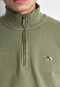 Olijfgroen half-zip pullover van zacht materiaal, met een gestructureerd oppervlak en een klein geborduurd logo op de linkerborst.