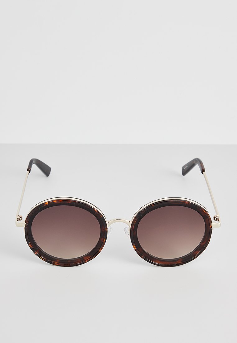 Occhiali da sole rotondi con montatura in tartaruga, lenti gradienti marroni, sottili dettagli in metallo dorato e punte nere sulle stanghette.