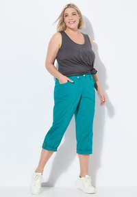 Pantaloni cropped color turchese con tasche laterali, orli arrotolati e bottoni metallici, abbinati a un top grigio senza maniche. Sneakers bianche completano l'outfit.