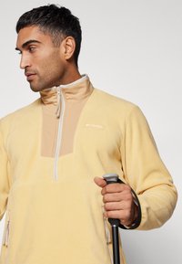 Fleece pulóver világos sárga színben, magas gallérral és fél cipzárral. Különböző bézs panelek és oldalsó zsebek jellemzik; sima textúra.