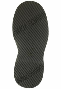 Suela de zapato de goma negra con surcos texturizados, con "STEVE MADDEN" grabado. Forma ligeramente redondeada con una parte inferior plana.