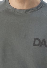 Detailní záběr na šedou mikinu s kulatým výstřihem, na prsou s částečně potisknutými černými písmeny "DA".