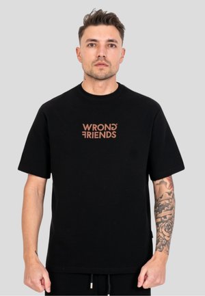 Mężczyzna z krótkimi włosami, ubrany w czarną koszulkę z brązowym napisem "WRONG FRIENDS" i widocznym kwiatowym tatuażem na prawym przedramieniu, stoi na szarym tle.