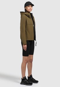 Giacca impermeabile verde oliva con cappuccio, zip nera, indossata su un top nero e pantaloni ciclistici, abbinata a scarpe da ginnastica nere e chunky.