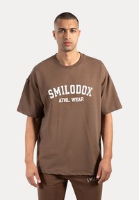 Camiseta oversized marrón hecha de una tela suave, con un logo blanco "SMILODOX ATHL. WEAR" impreso en el frente. Diseño simple con cuello redondo.
