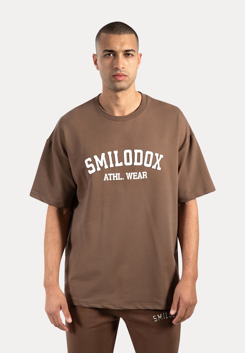 Camiseta oversized marrón hecha de una tela suave, con un logo blanco "SMILODOX ATHL. WEAR" impreso en el frente. Diseño simple con cuello redondo.