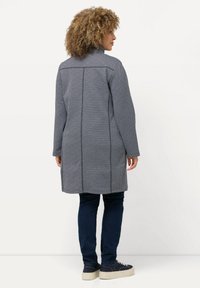 Cappotto grigio a fantasia con un design geometrico, maniche lunghe, colletto alto e taglio dritto, abbinato a jeans in denim scuro e sneakers.