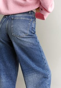 Persoană purtând blugi albaștri cu talie înaltă și croială lejeră și un pulover roz, pufos, văzută din spate, pe un fundal simplu, deschis la culoare.