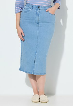 Mujer con falda midi vaquera azul claro, camiseta a rayas rosa y azul, y zapatos crema de tejido trenzado sin cordones, de pie con una mano en el bolsillo.