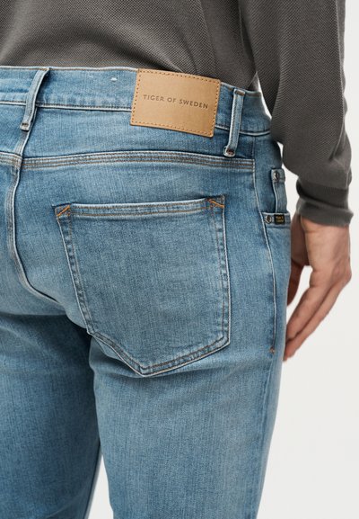 Άτομο που φοράει τζιν παντελόνι ανοιχτού μπλε χρώματος με πίσω τσέπη και δερμάτινη ταμπέλα καφέ χρώματος με την επιγραφή "Tiger of Sweden".