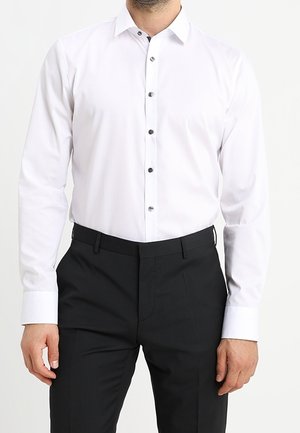 Homme portant une chemise blanche ajustée avec des boutons noirs, rentrée dans un pantalon habillé noir, sur un fond uni.