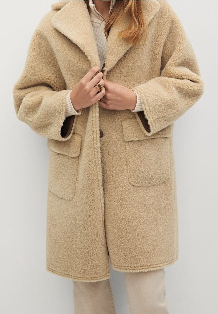 Manteau beige en peluche avec un large col, deux poches avant et une fermeture à boutons, fabriqué en matière polaire texturée.