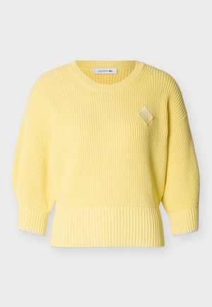 Rumena rebrasta pletena majica z okrolim izrezom, 3/4 balonastimi rokavi in majhno diamantno oblikovano nalepko na prsih.