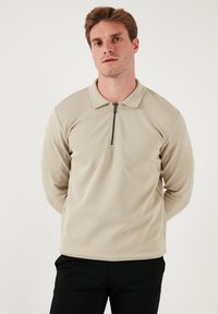 Polo beige a maniche lunghe con colletto zip, tessuto testurizzato e vestibilità aderente. Indossato con pantaloni neri, caratterizzati da una vestibilità rilassata.