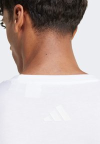 Biely tričko s okrúhlym výstrihom a textúrovaným logom Adidas z reflexného materiálu na hornej časti chrbta. Hladká tkanina, krátke rukávy.