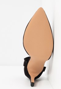 Chaussure à talon aigu noir en daim, photographiée par le dessous, montrant une semelle beige et un talon fin avec un embout en caoutchouc.