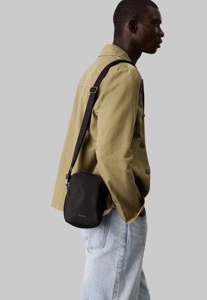 Sort rektangulær crossbody-taske med lynlåslukning, glat tekstur og justerbar rem, båret på hoften af en person i en grøn jakke.