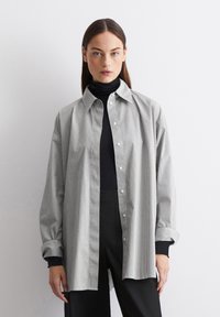 Chemise à rayures noire et blanche, avec un col et des manches longues à revers, portée par-dessus un col roulé noir.