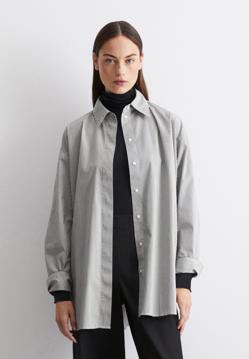 Chemise à rayures noire et blanche, avec un col et des manches longues à revers, portée par-dessus un col roulé noir.